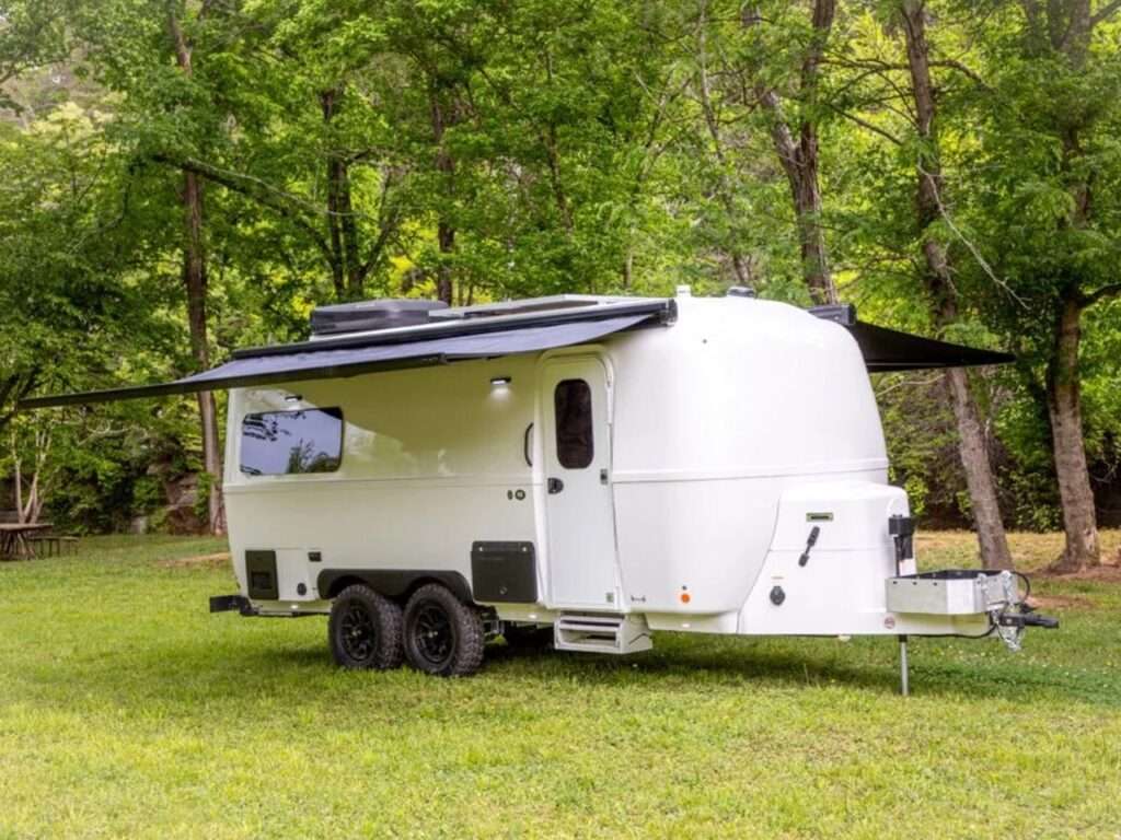 Side view of the Oliver Apex X23 fiberglass travel trailer with extended black awning in a lush green forest setting.