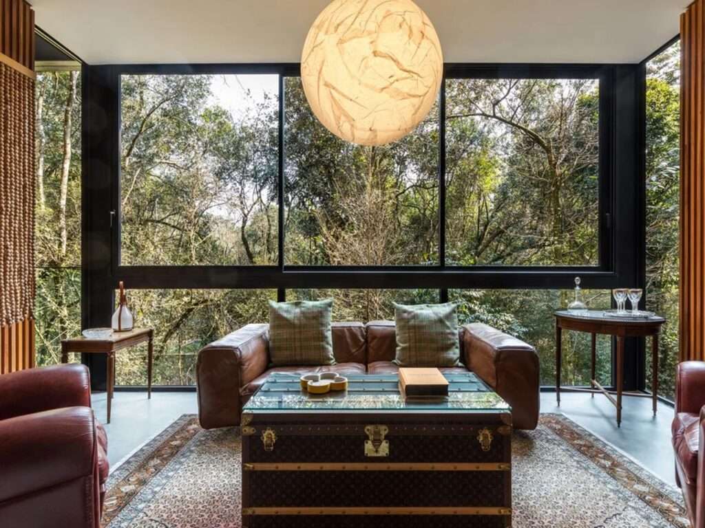 Sophisticated living room in House 17-JB featuring leather sofas, an antique trunk coffee table, and floor-to-ceiling windows overlooking the woods.