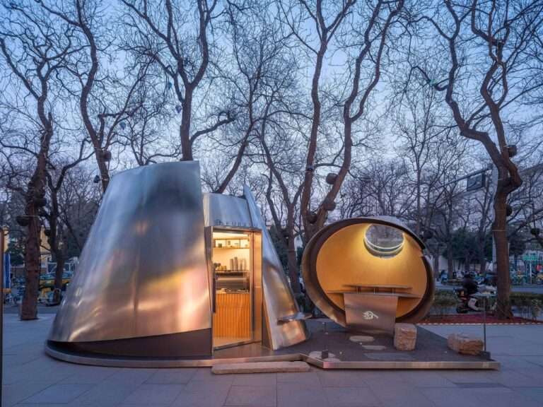 Exterior dusk view of the Kurasu coffee pop-up pavilion in Beijing, featuring two interlocking stainless steel geometric masses under bare trees.