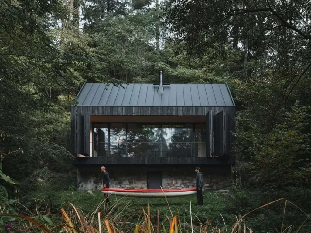 A dark charred wood cabin built on a historic stone base by a river, with two people carrying a red canoe in the foreground.