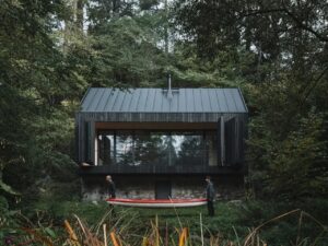 A dark charred wood cabin built on a historic stone base by a river, with two people carrying a red canoe in the foreground.