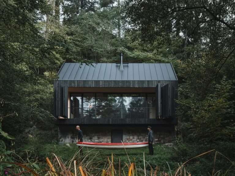 A dark charred wood cabin built on a historic stone base by a river, with two people carrying a red canoe in the foreground.