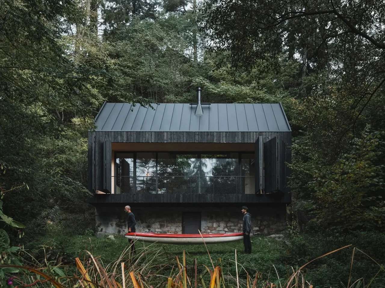 A dark charred wood cabin built on a historic stone base by a river, with two people carrying a red canoe in the foreground.