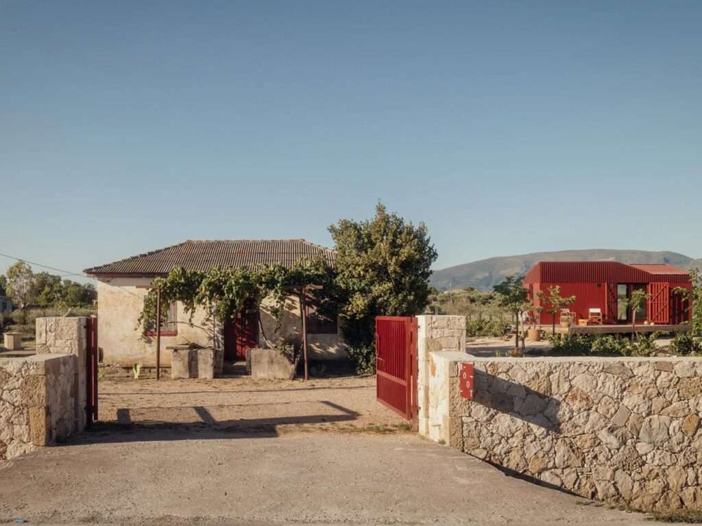 A historical traditional plaster house next to the modern prefab deep red Root Cabin by Kasawoo behind a stone wall entrance in Zakynthos.