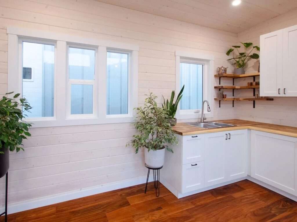 Modern open-plan kitchen inside a tiny home featuring white cabinetry, warm butcher block countertops, wooden floating shelves, and indoor potted plants.