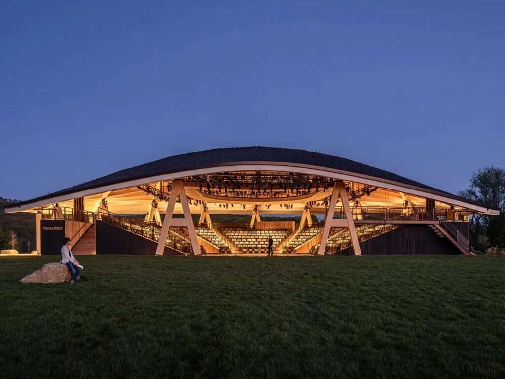 Illuminated open-air amphitheater of the Hudson Valley Shakespeare permanent facility by Studio Gang at dusk.