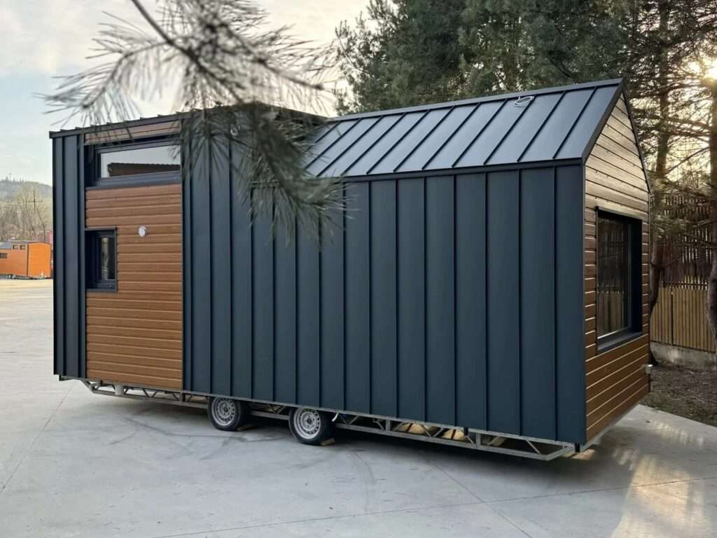 Side profile of the Smile mobile home on a steel trailer with wheels, showing the dark metal roofing and wood accents.