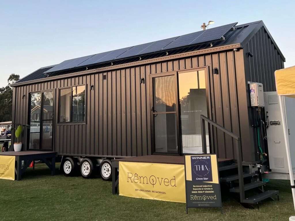 Exterior view of Tallebudgera 9.6 tiny home by Removed Tiny Homes, featuring dark gray Colorbond steel cladding, a triple-axle trailer, and rooftop solar panels at an outdoor expo.