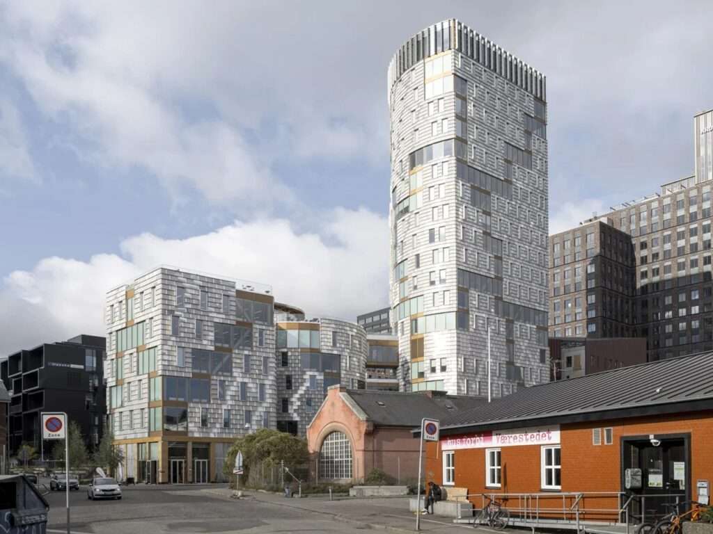 Street-level view of the TRAE building in Aarhus showcasing the contrast between the timber high-rise and existing low-rise neighborhood structures under a cloudy sky.