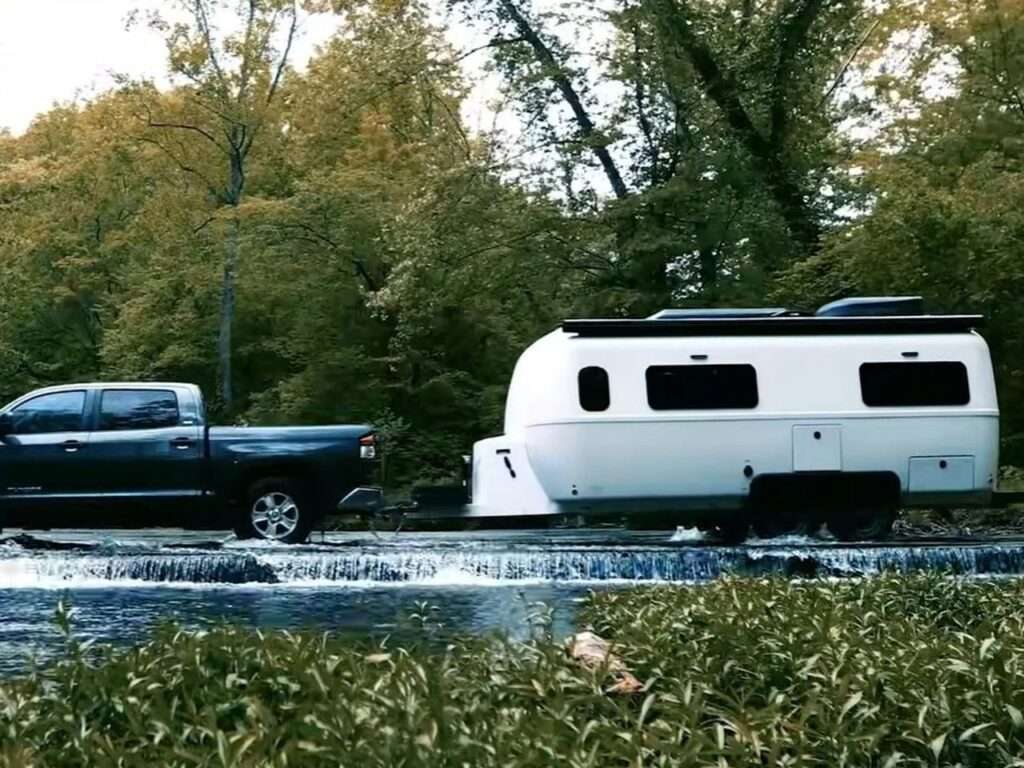 A dark pickup truck towing the Oliver Apex X23 travel trailer through a shallow river crossing in a wooded area.