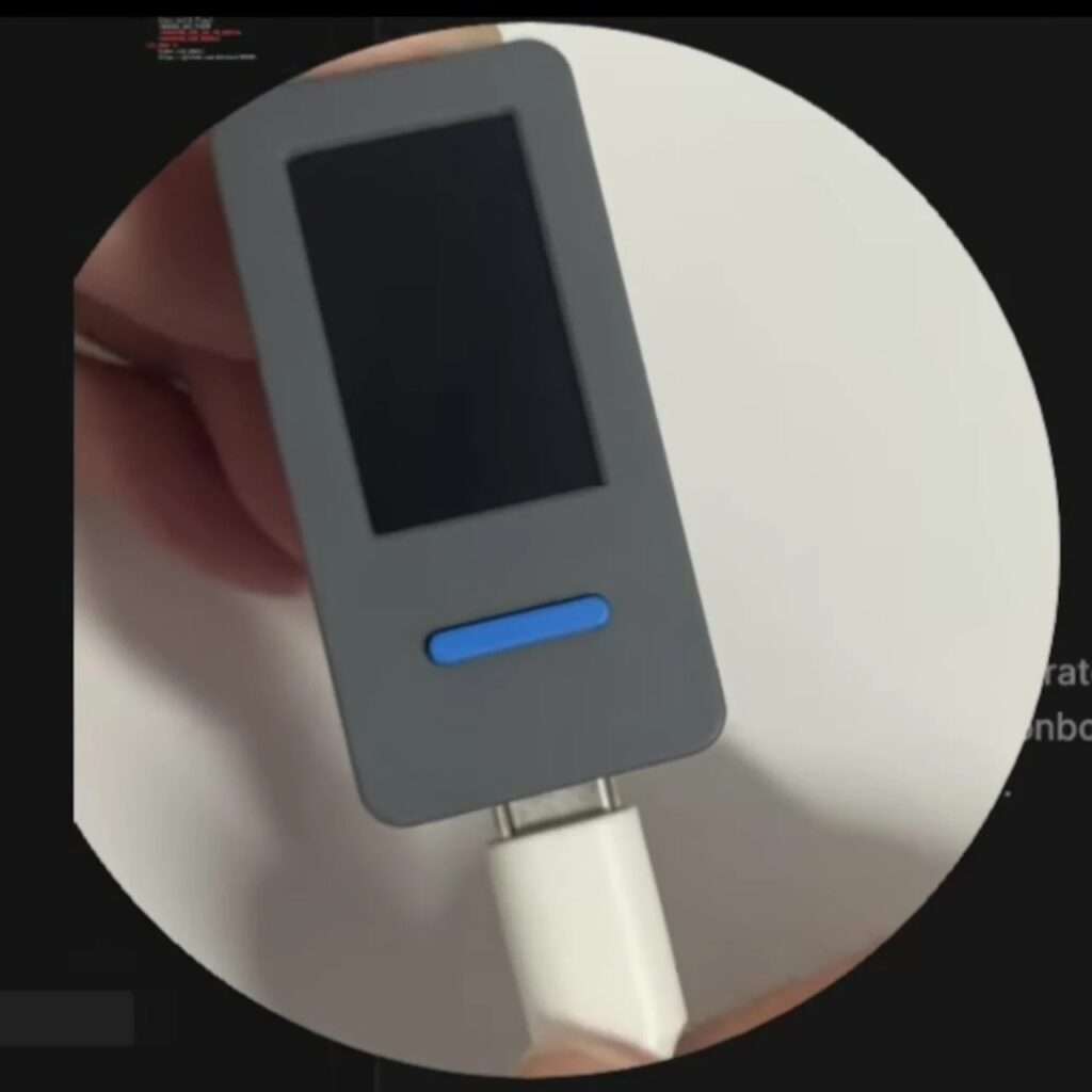 A hand holding a grey compact hardware stick with a blue horizontal button while a white USB Type-C power and data cable is being connected to its bottom port.
