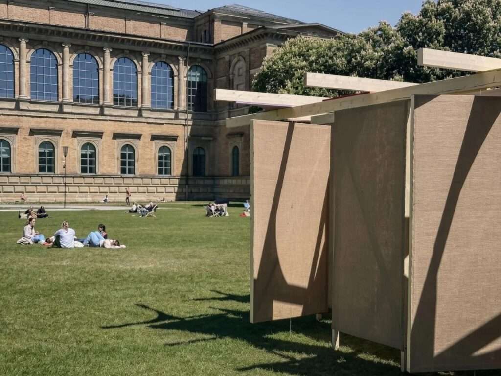 Angled view of the DELULU pavilion's raw jute panels in the foreground, with visitors relaxing on the grass field of the historic Alte Pinakothek museum building under a clear blue sky.