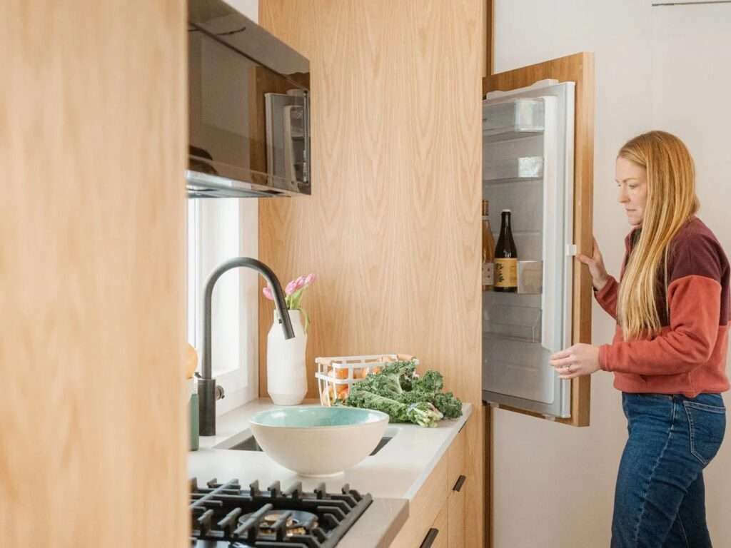 A woman opening an integrated wood-paneled refrigerator inside the compact kitchen of the Halcyon Grand tiny home.