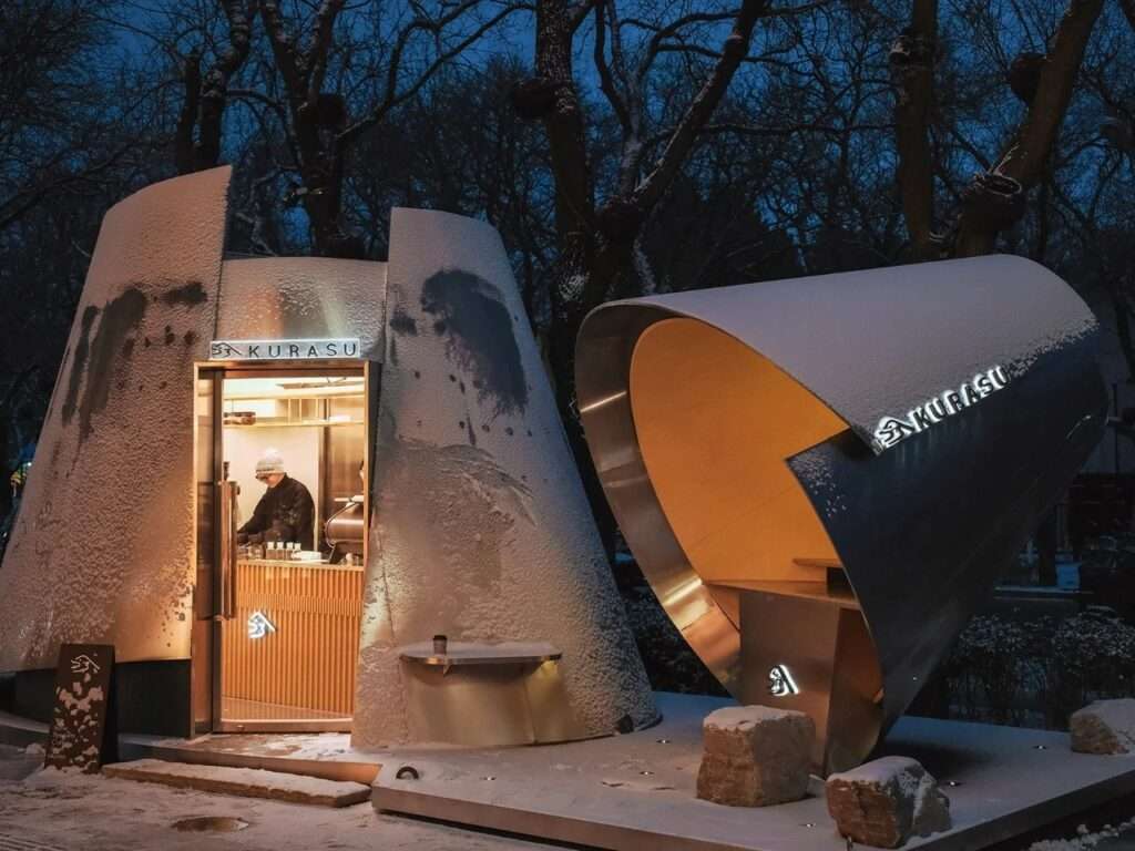 Snow-covered Kurasu coffee pavilion in Beijing at night, showing illuminated warm wood interior and glowing brand signage.