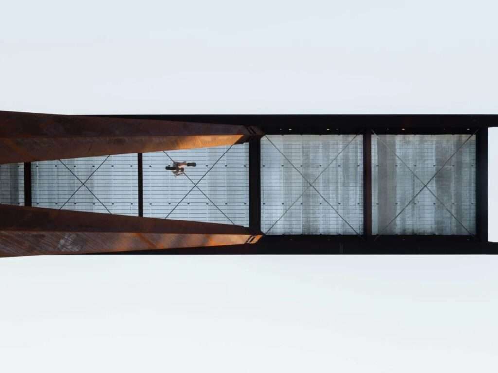 Underside view of a steel mesh floor lookout platform showing structural support beams and a visitor walking above.