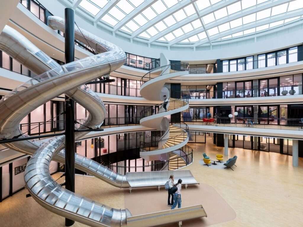 Interior of the Philo Center at Institut Le Rosey featuring a vast central atrium under a glass skylight with a multi-story spiraling stainless steel slide and a white concrete spiral staircase.