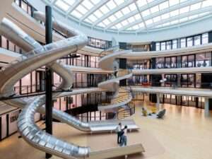 Interior of the Philo Center at Institut Le Rosey featuring a vast central atrium under a glass skylight with a multi-story spiraling stainless steel slide and a white concrete spiral staircase.