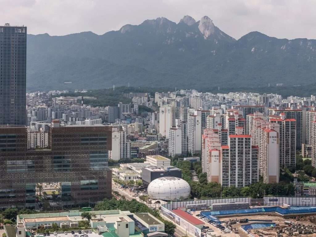 Distant aerial drone view of the spherical Seoul Robot and AI Museum building surrounded by apartment complexes with a massive mountain range in the background.