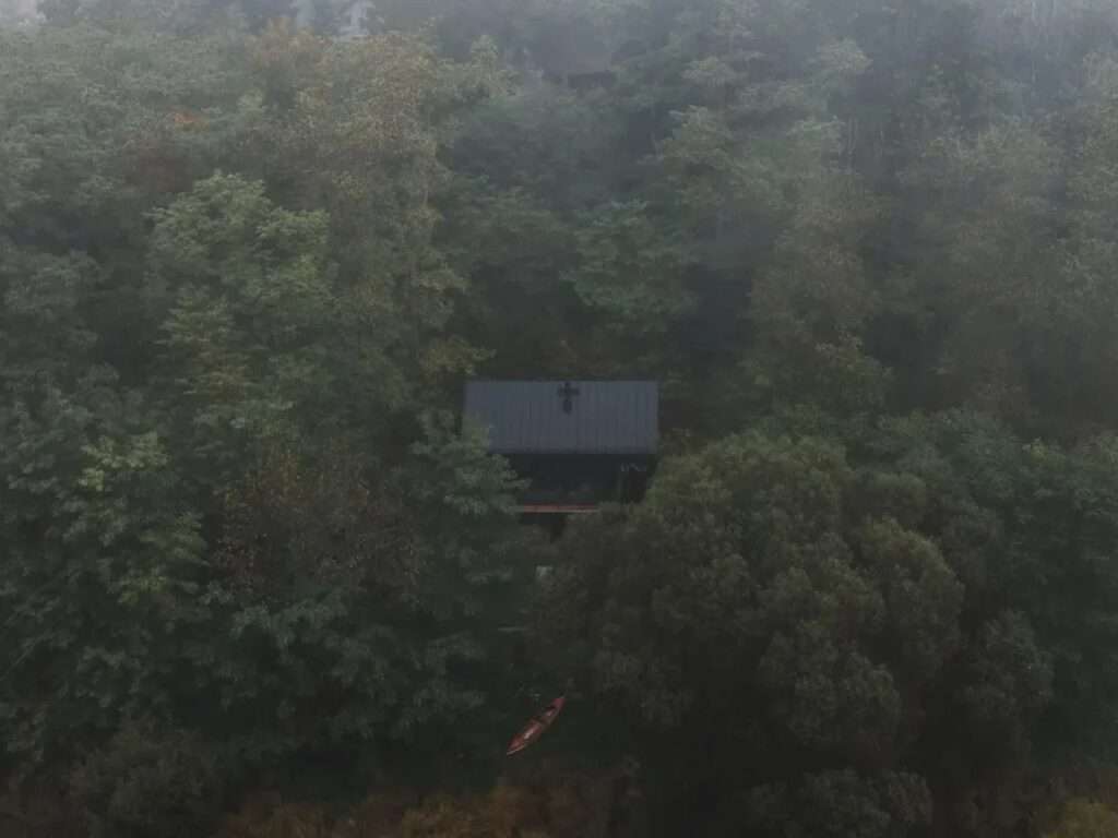 Aerial view of a dark minimalist cabin camouflaged within a dense green pine forest under misty weather.