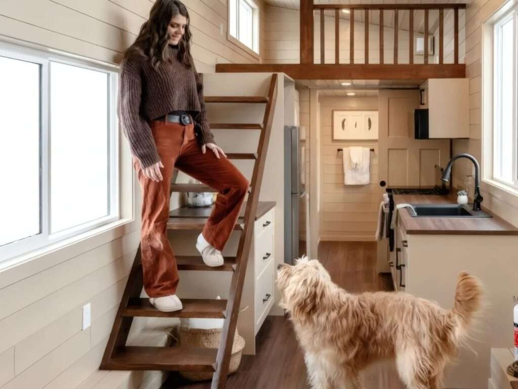 A woman stepping down a custom wooden ship-ladder in the kitchen area of the Ruby tiny home, with a golden dog looking up from the hardwood floor.