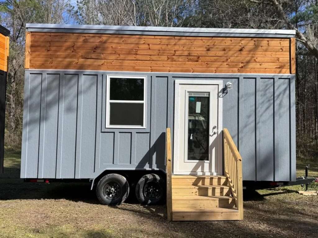 Exterior view of the Shanty tiny house featuring gray board-and-batten siding, natural wood upper trim, and a small wooden staircase leading to the white front door.