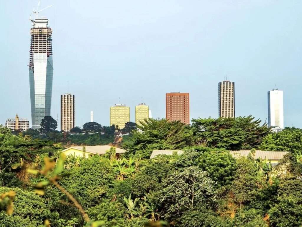 Distant landscape view of Tour F Tower under construction in Abidjan, showing its growing concrete core emerging high above trees and older administrative towers.