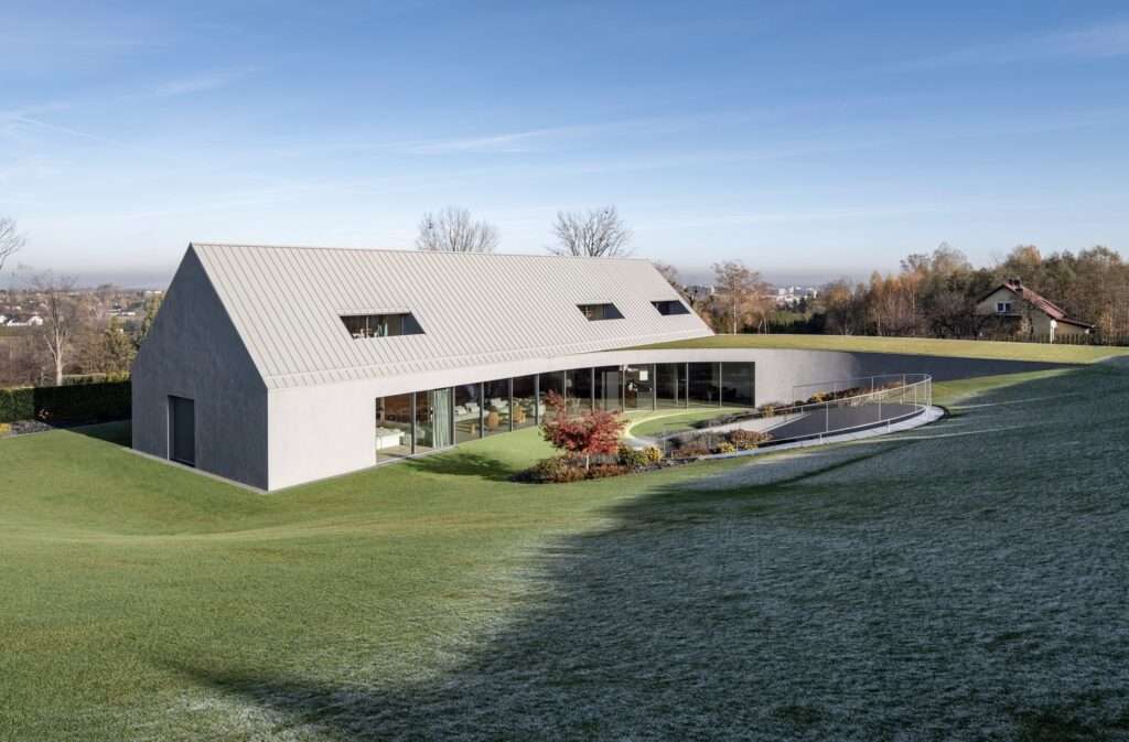 Rear garden elevation of the Yaw House featuring a long glass facade, an upper gabled structure, and a circular ramp leading into a subterranean courtyard.