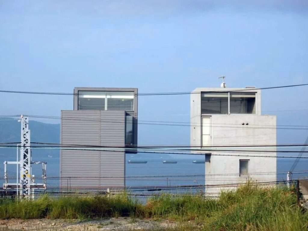 Distant coastal view of Tadao Ando's twin 4x4 Houses behind overhead utility lines, overlooking the Seto Inland Sea with ships on the horizon.