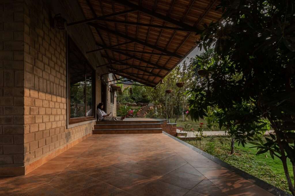 A woman sitting on a wide shaded veranda floor paved with terracotta tiles under a deep sloping roof structure next to a compressed earth block wall.