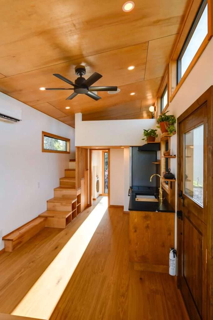 Full-length interior view of the Black Butte tiny home showing the open-plan layout, wood ceilings, storage stairs, kitchen counter, and loft joists.