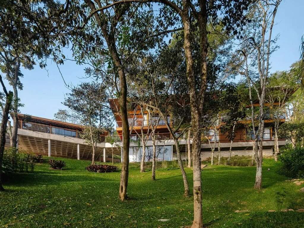 Wide shot of Casa Pinhal nestled within dense native trees of the Mantiqueira Mountains under a clear blue sky.