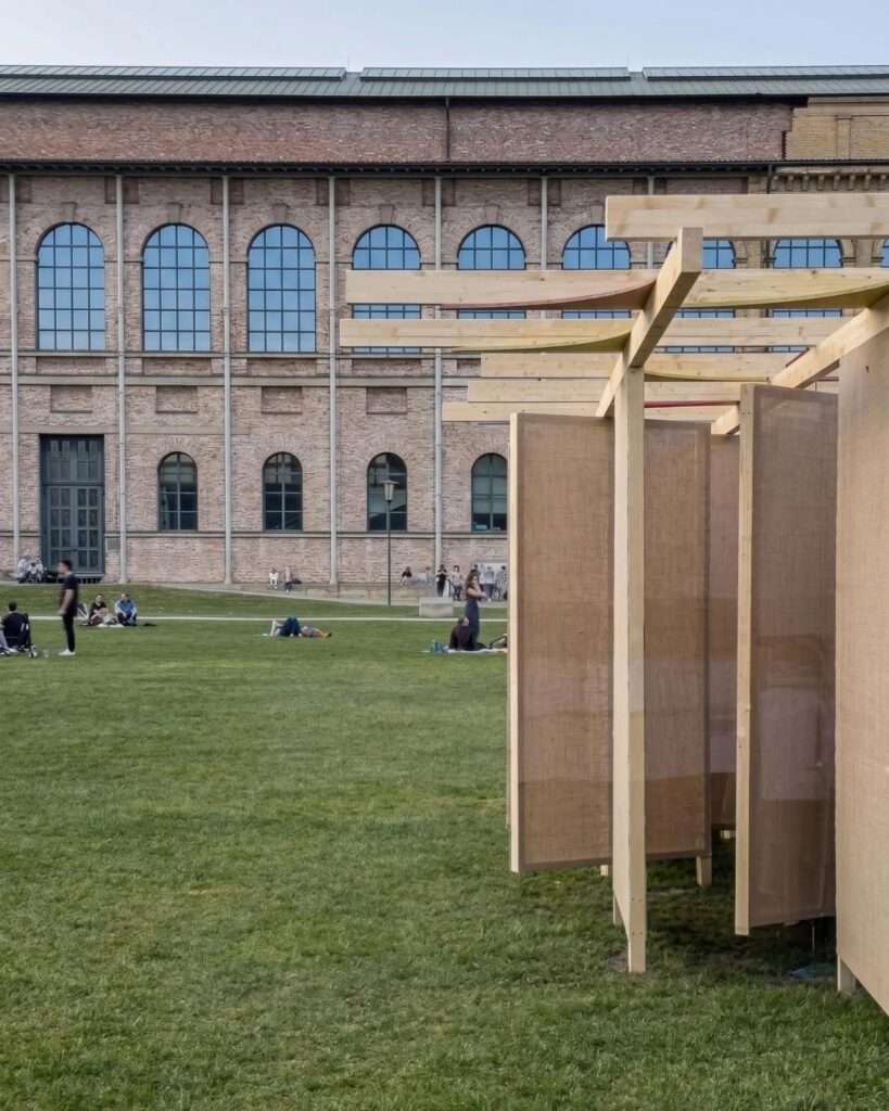 Side view of the linear timber beams and textured canvas walls of the DELULU installation, set against the grand arched windows of the Alte Pinakothek brick facade.