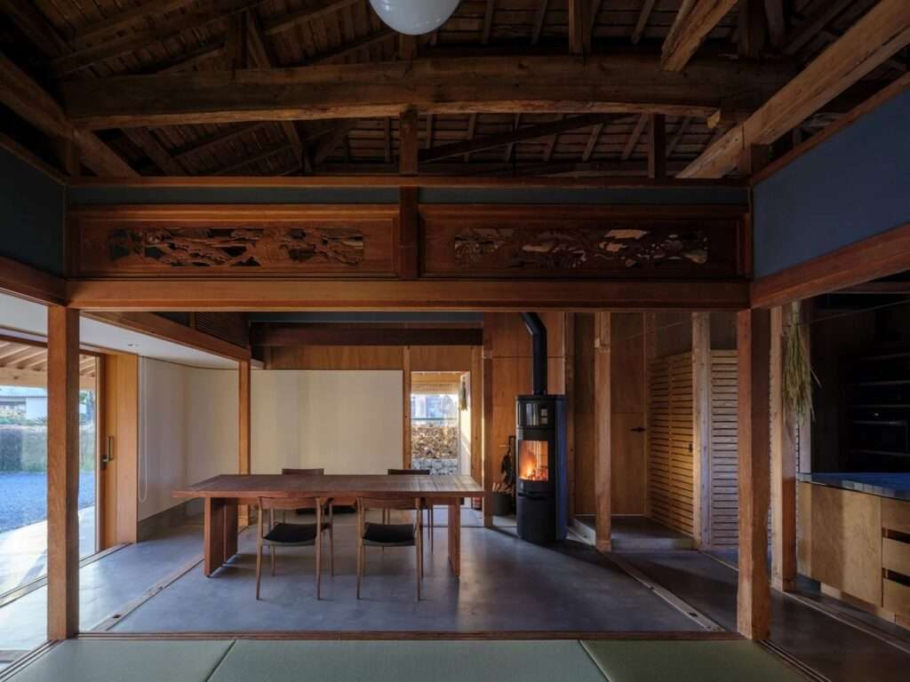 Open plan interior of a traditional Japanese house with exposed timber roof trusses, a wooden dining table, and a modern black freestanding fireplace stove.