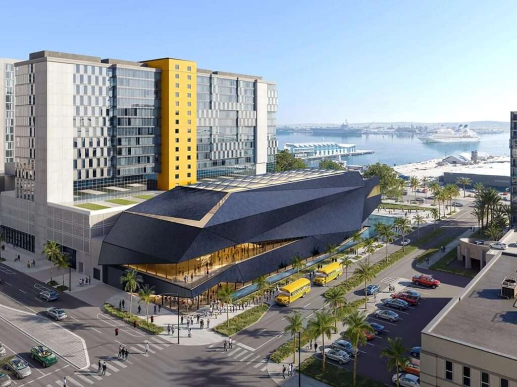 Aerial rendering of the proposed four-story Navy SEAL Museum San Diego building next to a high-rise tower, situated near Pacific Highway and the Embarcadero waterfront with naval vessels and cruise ships in the background bay.