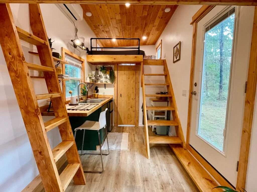 Interior walkway of Rasa tiny home with two wooden ladders leading to separate sleeping lofts and a central kitchen area.
