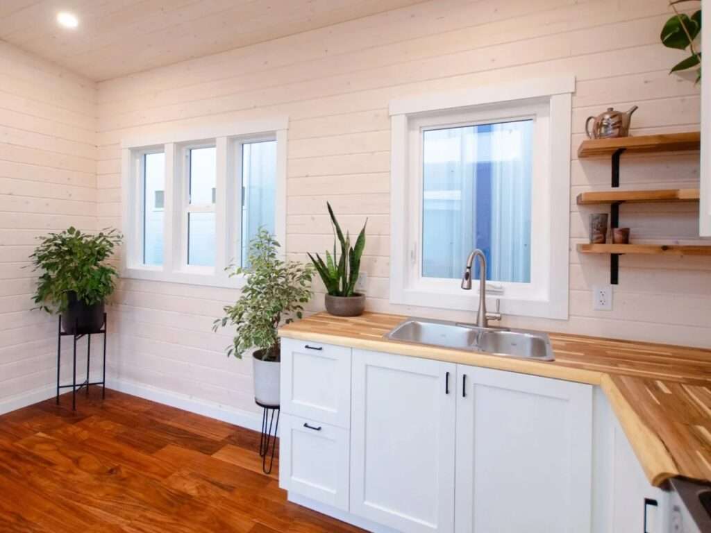 Close-up of a double-bowl stainless steel kitchen sink set in a light butcher block counter with white cabinets and floating wooden open shelves in a tiny house.