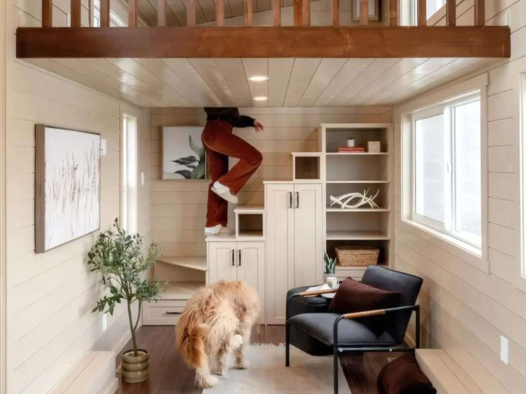 A person climbing a integrated storage staircase with built-in white cabinets and shelving units inside a tiny home living room.