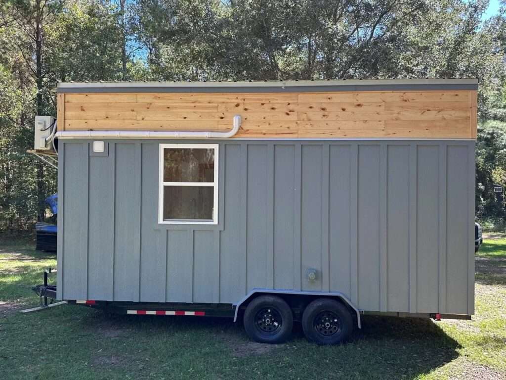 Side profile of the Shanty tiny house on a black steel trailer with dual wheels, showing the gray siding and upper wood accents against a forest background.