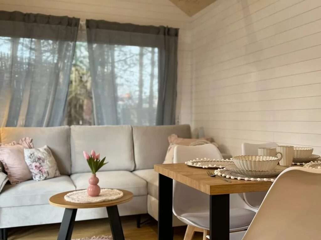Interior of the Smile tiny house living room with a grey sofa, wooden dining table, and large window with sheer curtains.