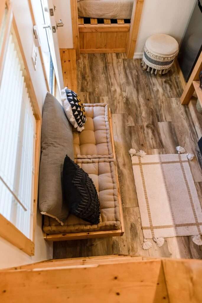 Top-down interior view from the loft looking down at the compact living room floor, built-in sofa cushions, and patterned rug of the Tulsi tiny home.