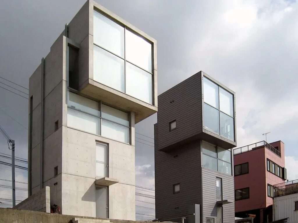 Three-quarter perspective angle of the two 4x4 Houses by Tadao Ando, emphasizing the vertical concrete and dark wood modular blocks under a cloudy sky.