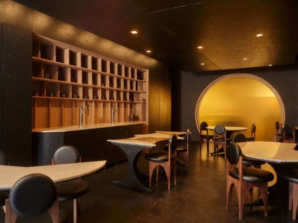 Wide angle view of Gourmega restaurant interior in Manhattan showing modular travertine tables, dark cork flooring, and the yellow kitchen portal.