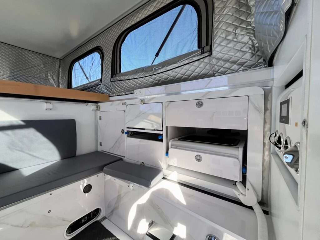 A side-angle interior view of the Roam Rider SL camper cabin, showcasing the convertible grey bench seat and the extensive white marble-patterned modular cabinetry system with sliding storage compartments.