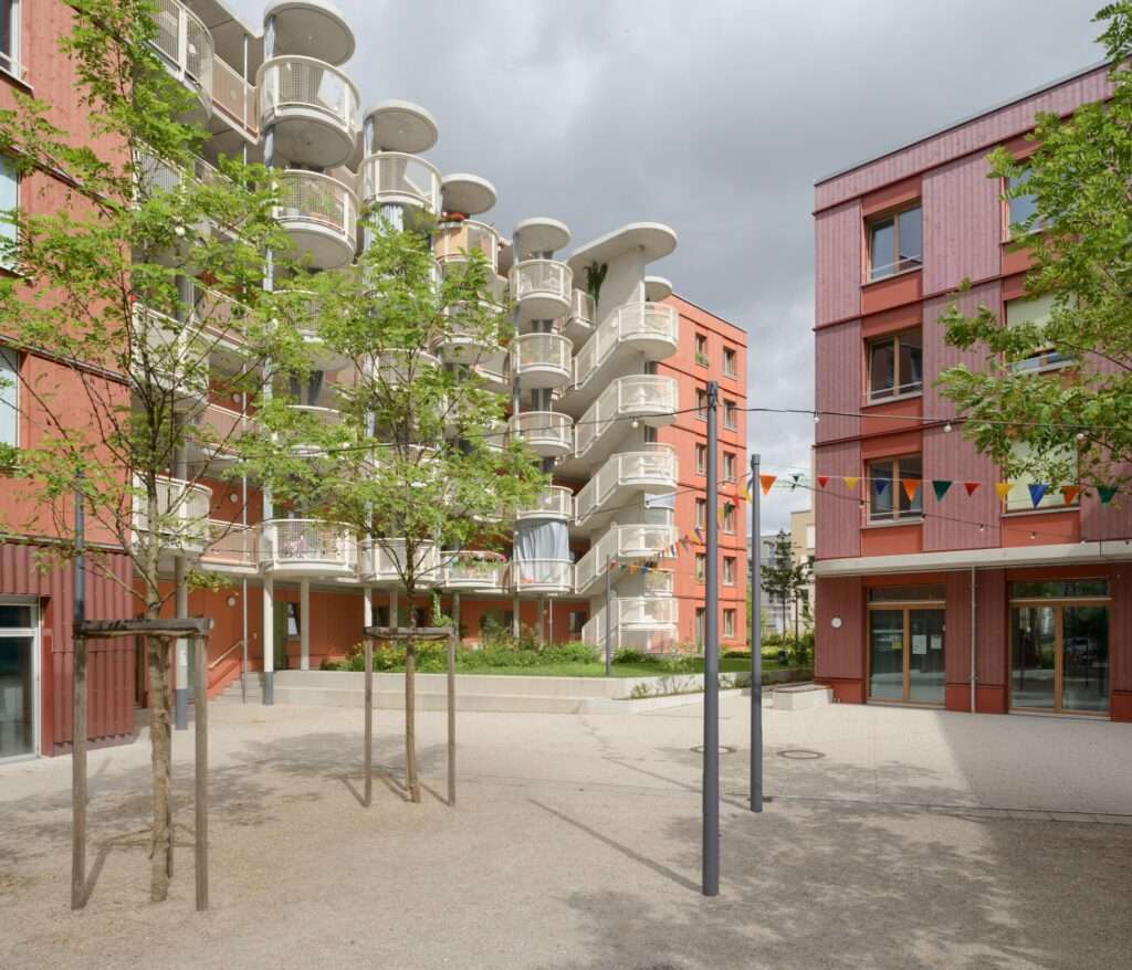 Open inner courtyard square with sand play area, young trees, and balconies of a cooperative housing block in Munich.