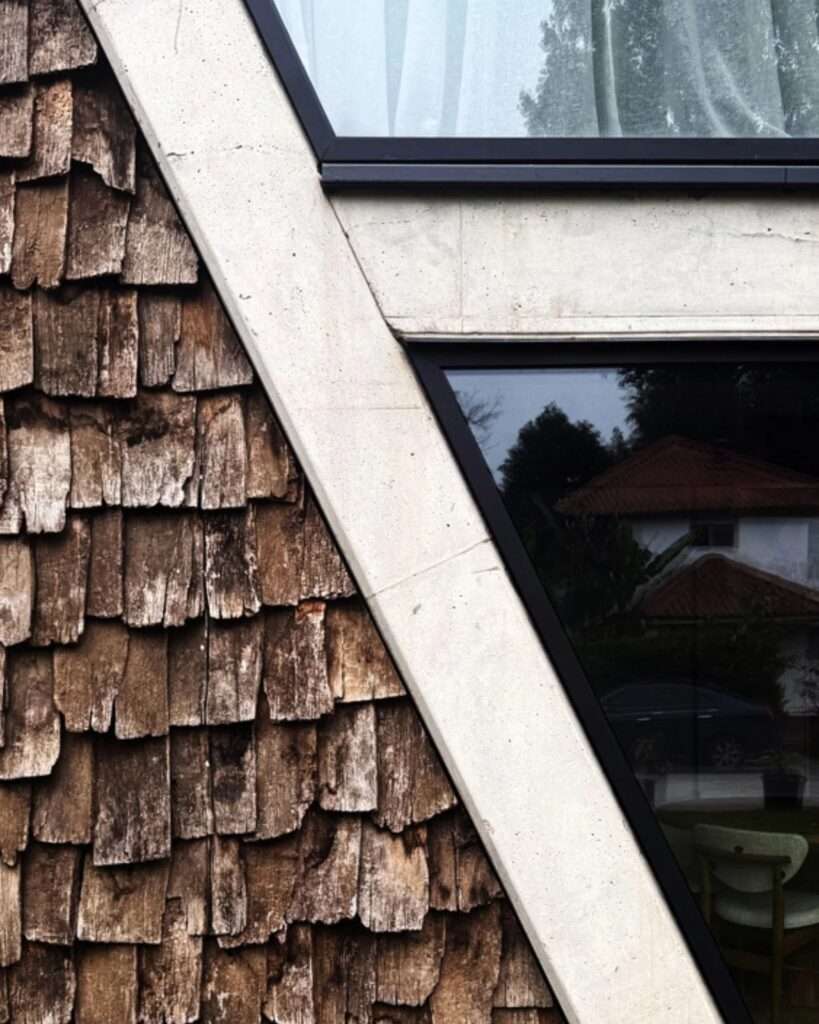 Macro detail of weathered wooden shingles meeting a crisp, angled raw concrete beam and reflective window glass.