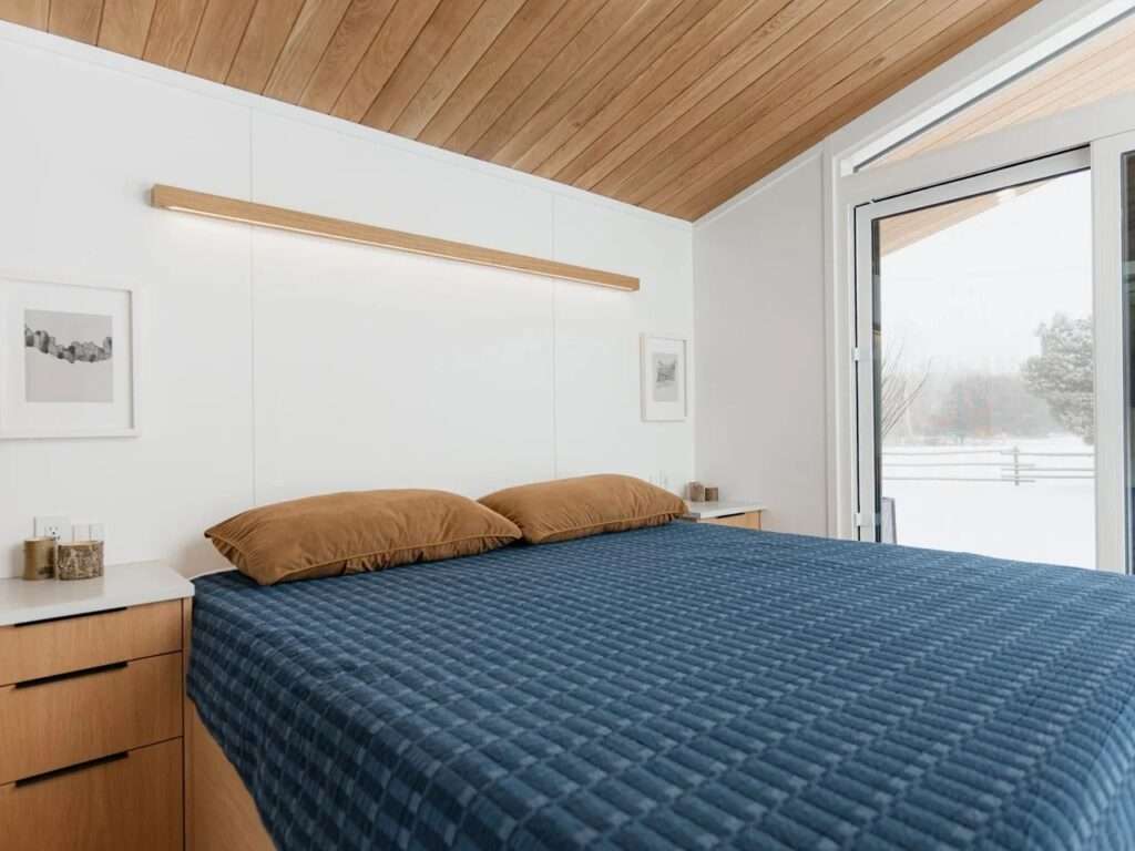 A large bed with a blue textured quilt and brown pillows in the main bedroom of the Halcyon Grand, looking out through sliding glass doors to a snowy view.