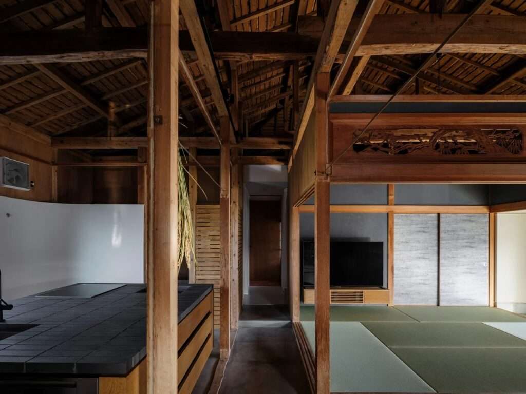 Interior view looking from a dark tile-clad kitchen island toward a traditional Japanese tatami room with sliding screens under an open wooden ceiling structure.