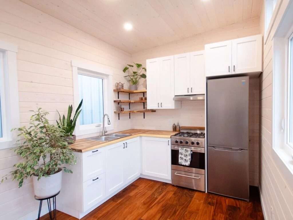 Compact kitchen design featuring a stainless steel gas stove, a full-size refrigerator, white upper cabinets, and a warm hardwood floor in a modern tiny home.