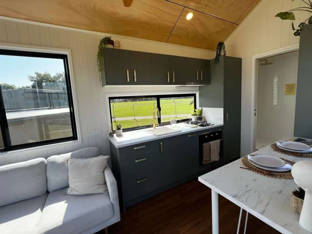 Compact interior living space of Tallebudgera 9.6 showing a modern gray fabric sofa, a round woven coffee table, a wall-mounted TV, and large panoramic windows with sunlight.