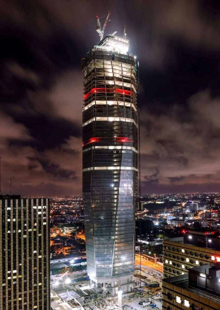 Nighttime drone photograph of Tour F Tower under construction, highlighting the semi-reflective glass facade panel installation and active tower cranes lit up against a cloudy night sky.